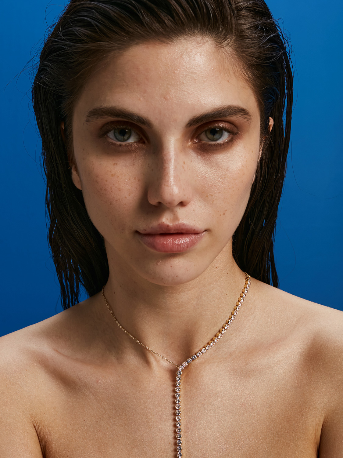 Before retouch — beauty close-up, freckled model with wet dark hair, blue background, diamond Y-necklace