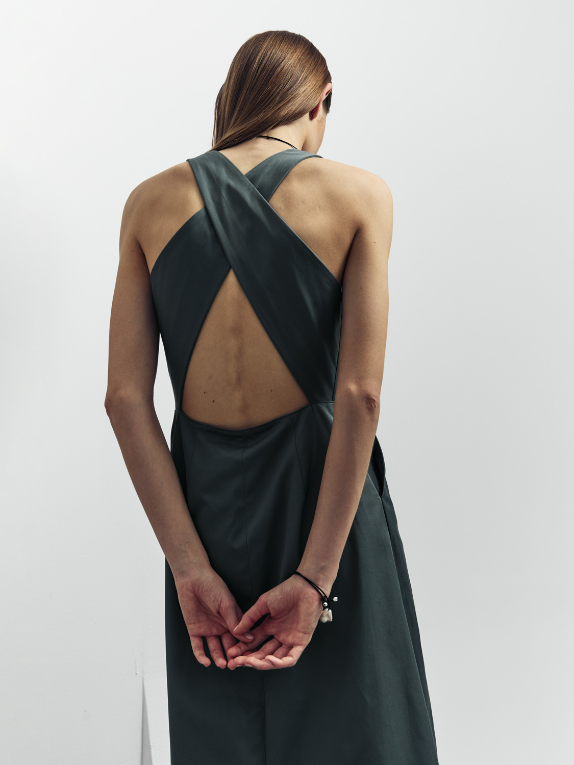 Before retouch — model in dark green crossback open-back dress, back view, light background, skin blemishes visible