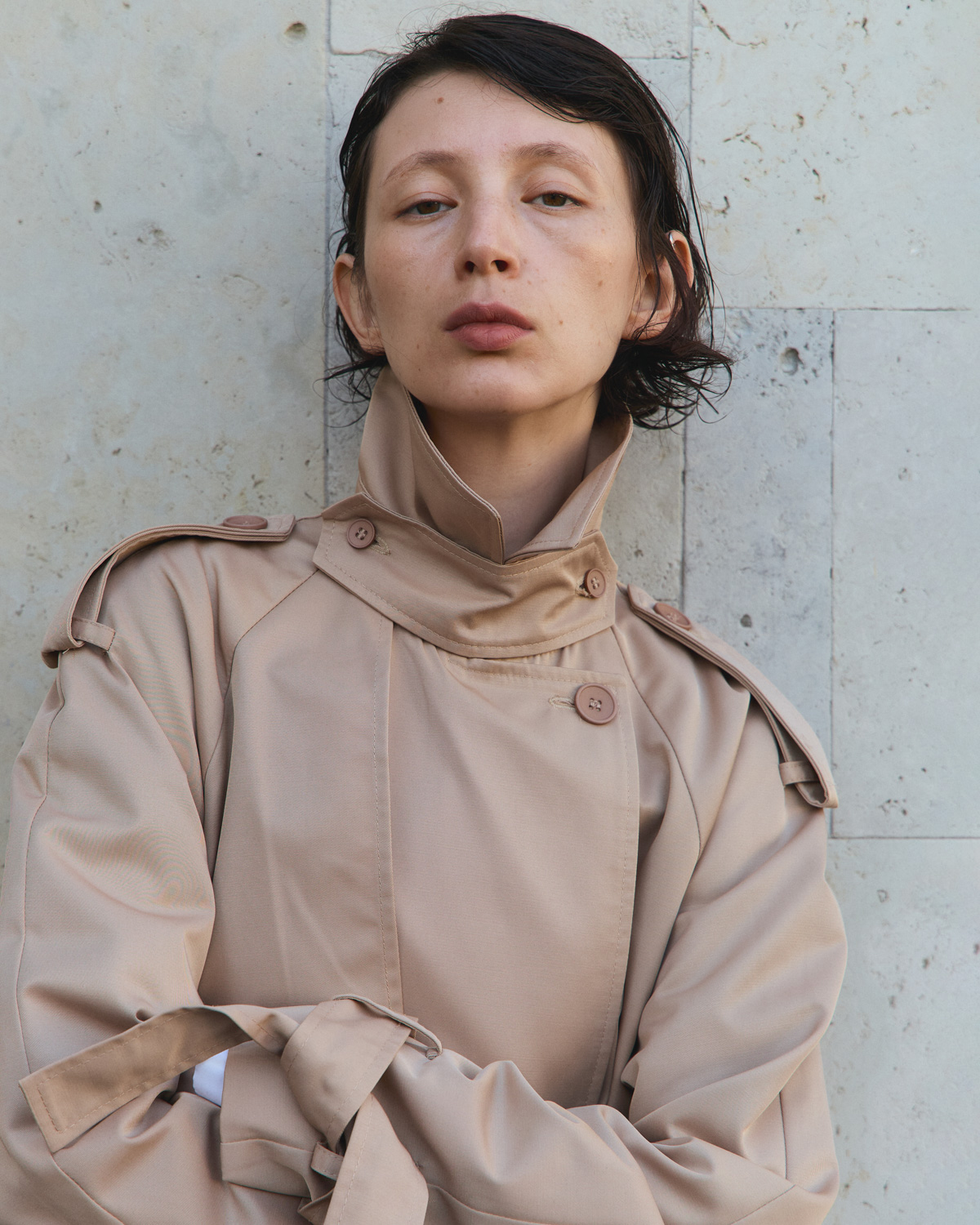 Before retouch — model in beige trench coat against travertine wall, visible skin blemishes and redness