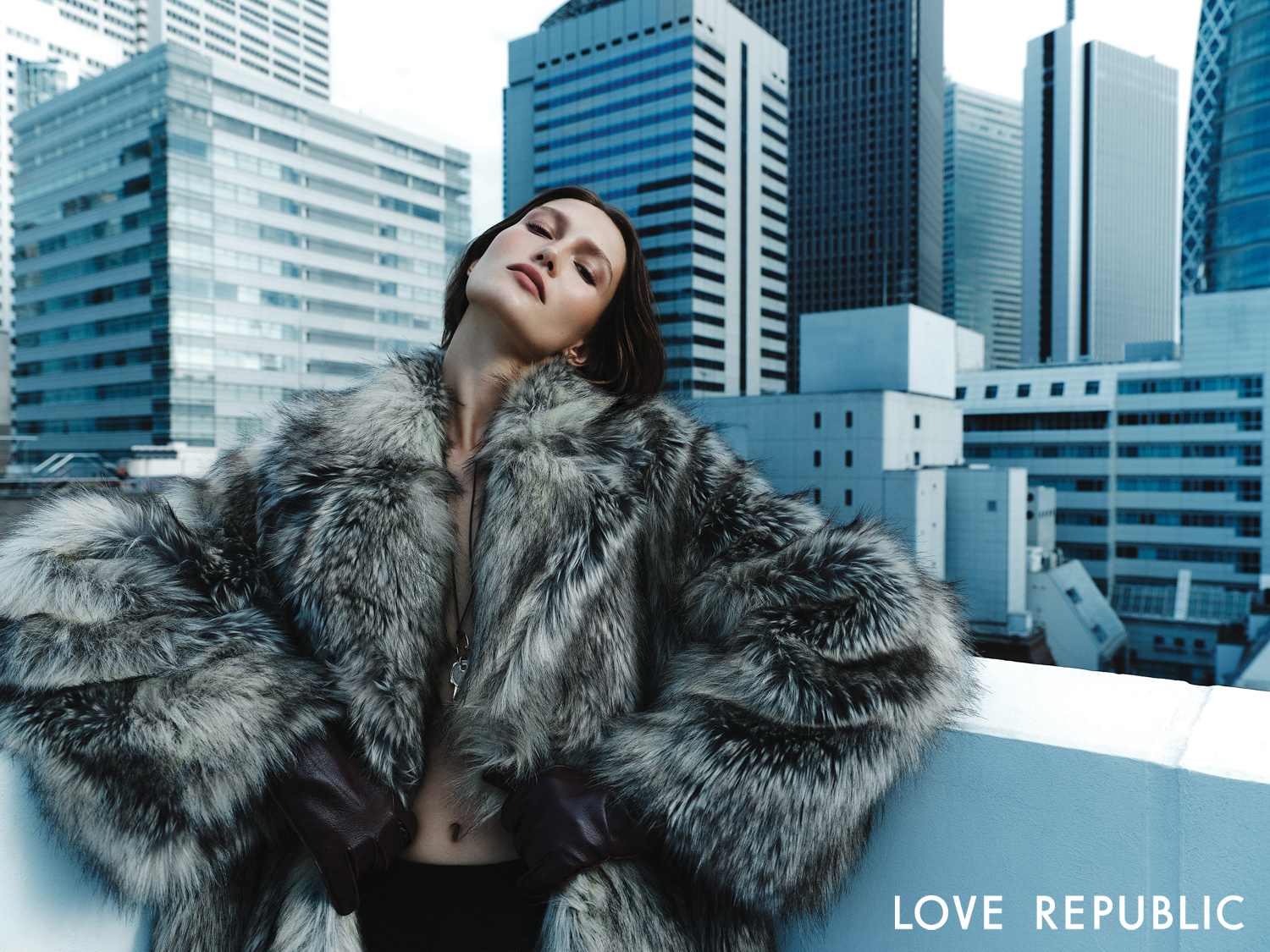 Editorial fashion portrait of a woman in a grey fur coat against a blue city skyline