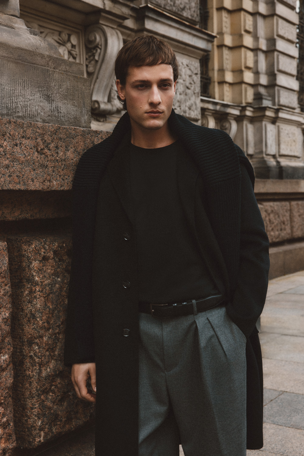 All We Need Campaign — male model in black ribbed sweater and grey trousers against ornate stone architecture