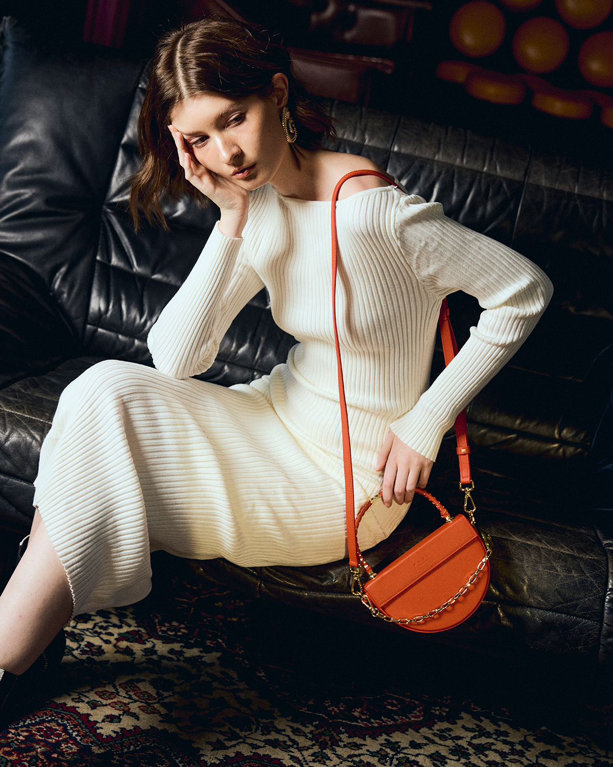Before retouch — woman in cream ribbed sweater on black leather couch with burgundy bokeh lights background, orange crossbody bag, natural raw state