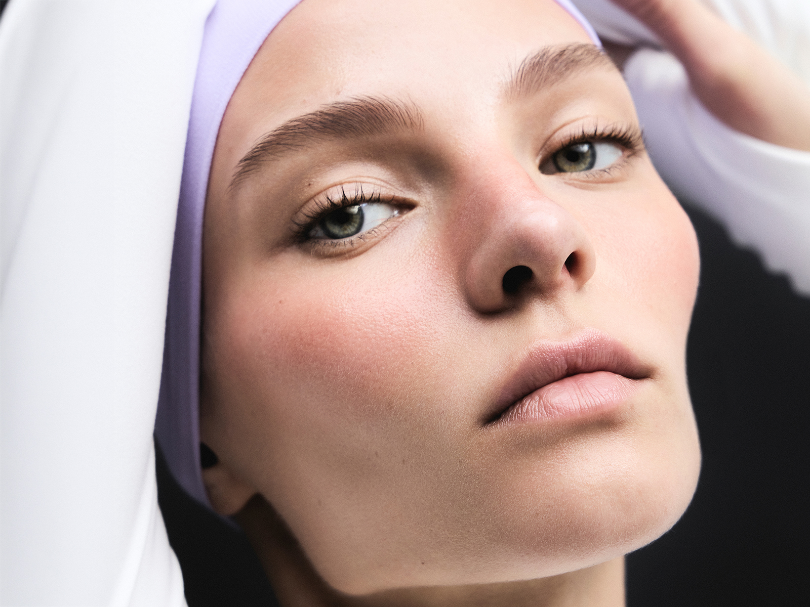 Close-up beauty portrait with green eyes, lavender headband, and luminous skin against dark background